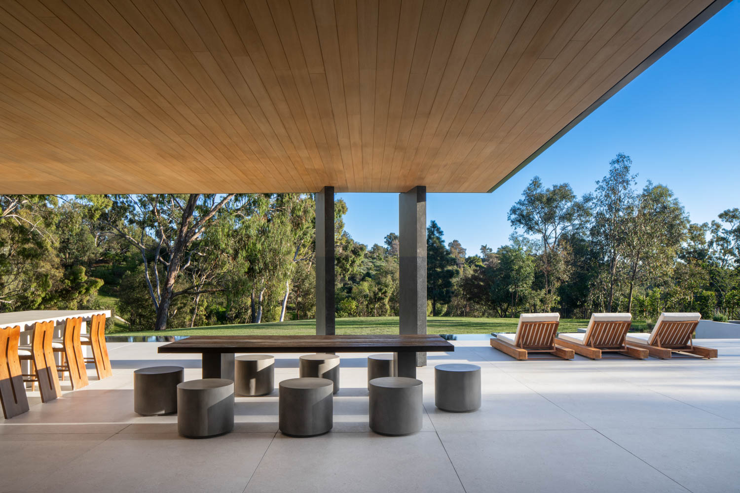 Modern outdoor patio with a long dining table, round stools, bar seating, and lounge chairs, set under a wooden ceiling with views of trees and a lawn.