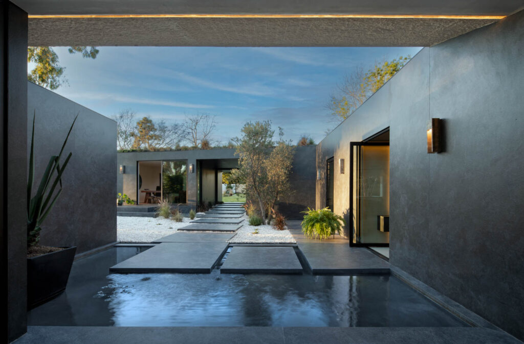 Modern courtyard with concrete stepping stones over a water feature, surrounded by minimalist architecture, plants, and open passageways to outdoor garden and living spaces.