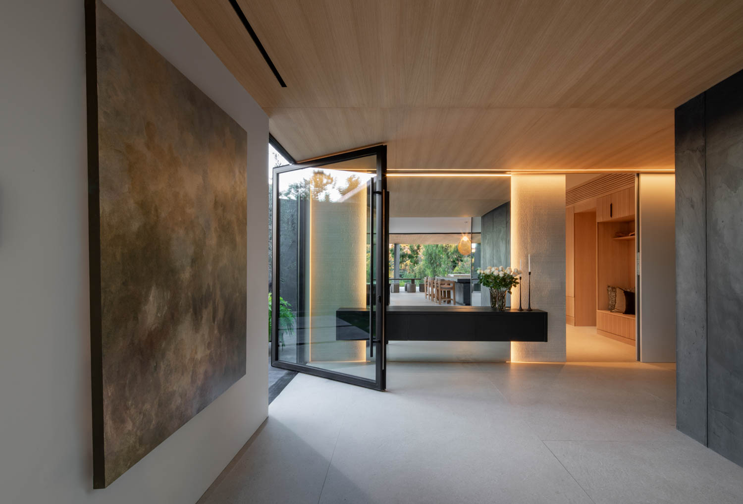 Modern entryway featuring a large abstract painting, glass door, minimalist decor, built-in bench, and warm recessed lighting leading to an open dining area.