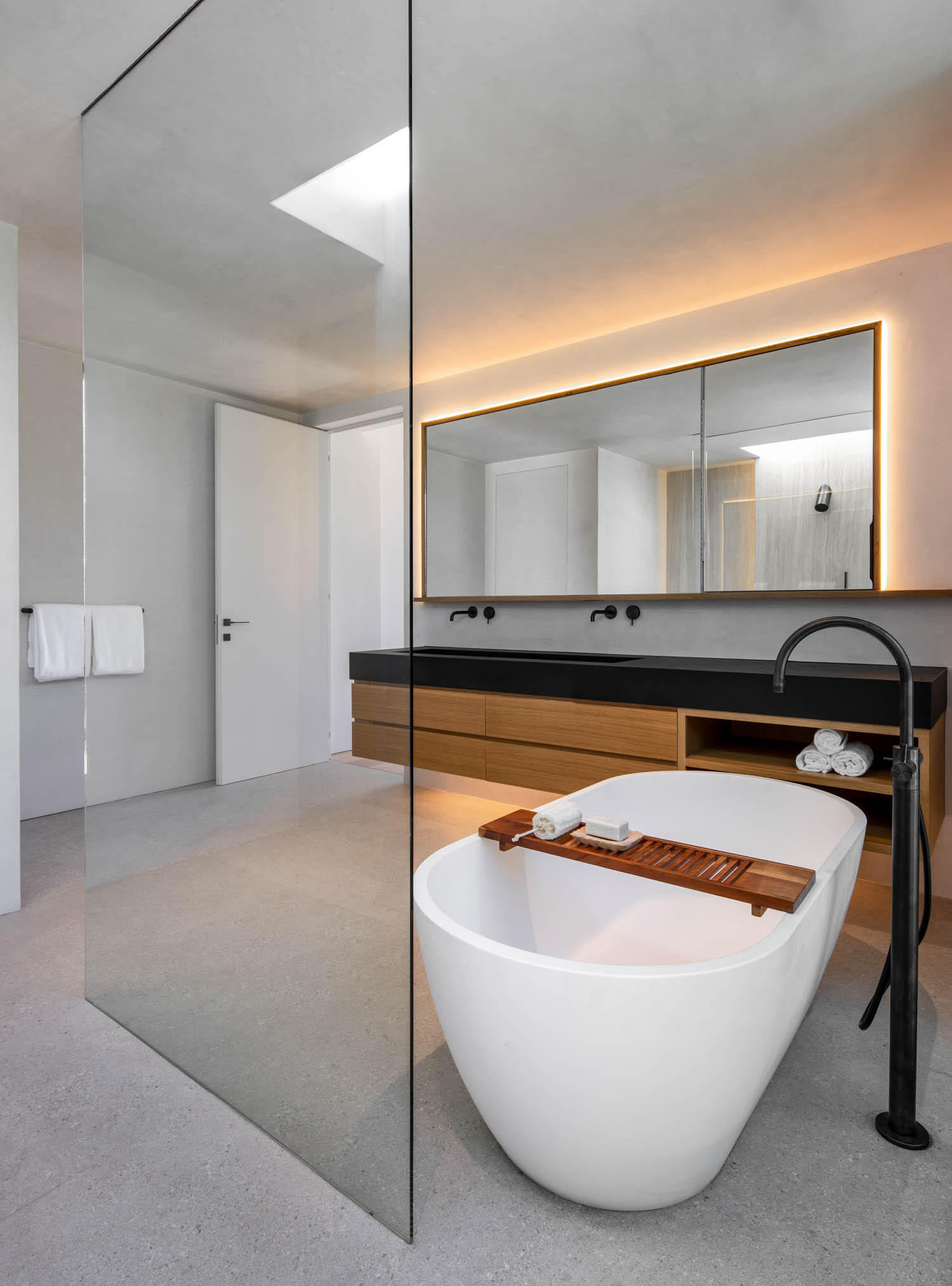 Modern bathroom with a freestanding white bathtub, wall-mounted black faucets, large mirror with backlighting, and a wooden bath tray; minimalist decor and neutral colors.