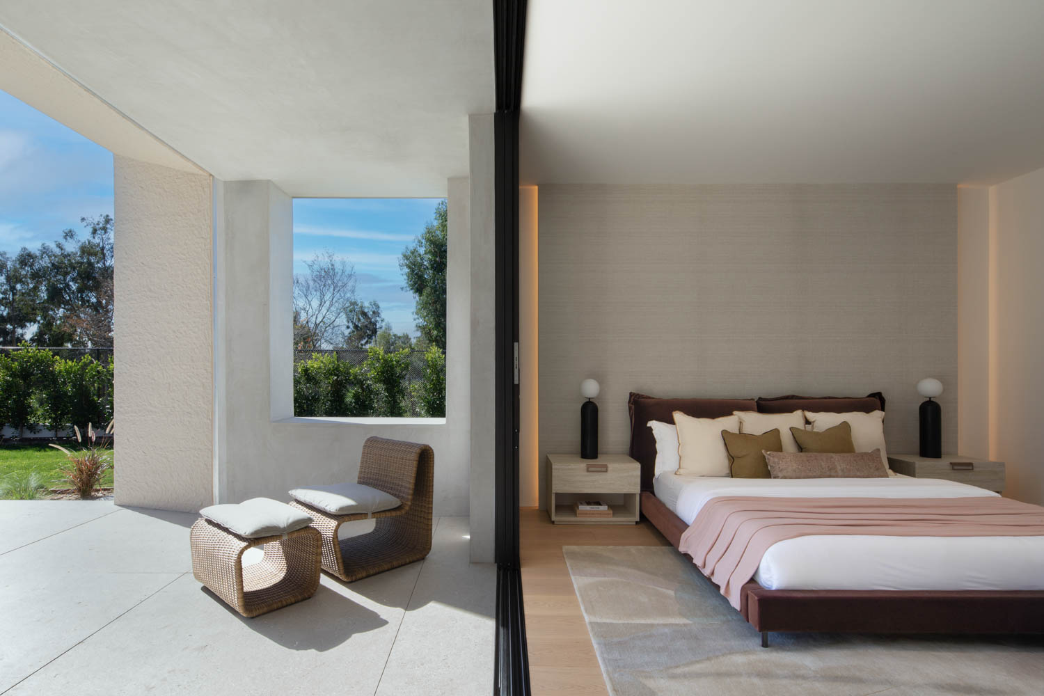 A modern bedroom with neutral tones opens to a patio with two wicker chairs and a small table, featuring large windows and abundant natural light.