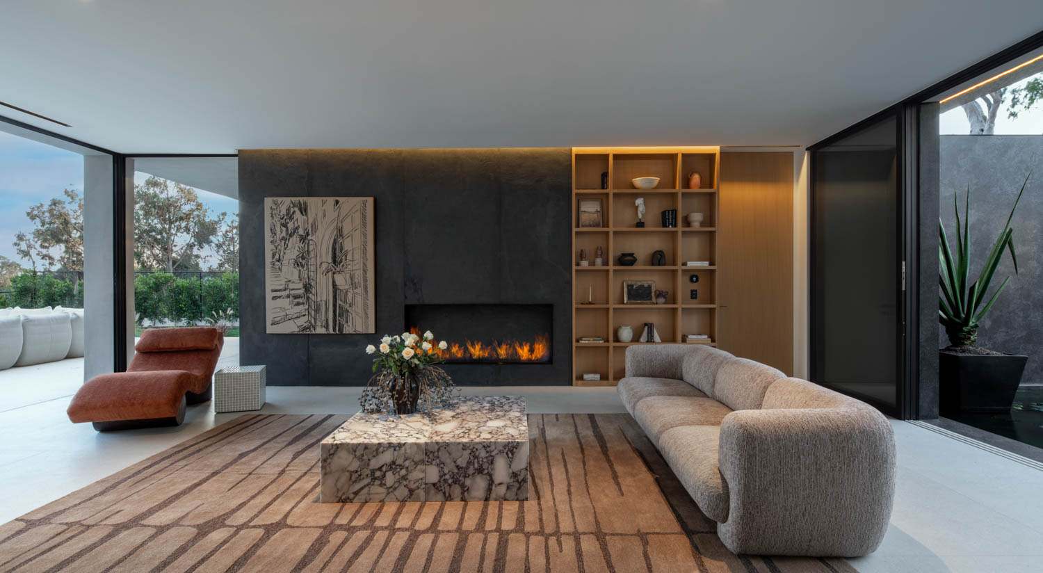 Modern living room with marble coffee table, beige sofa, brown lounge chair, built-in shelving, wall art, fireplace, and large glass doors opening to patio.
