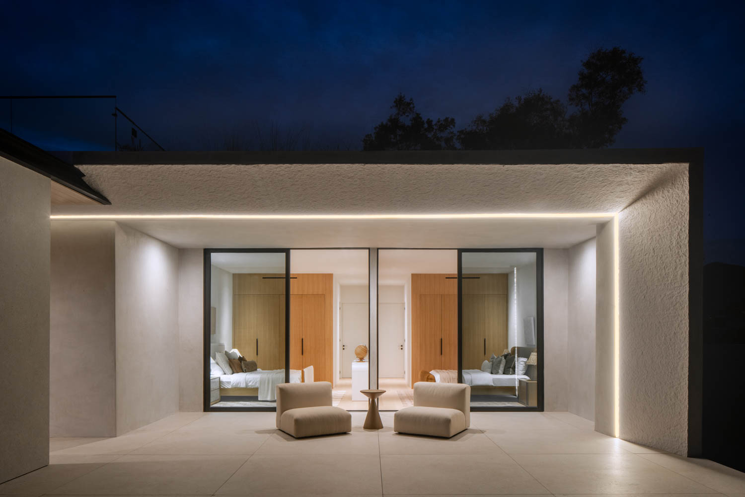 Modern patio at night with three white chairs and a small round table, facing large glass doors that reveal a well-lit interior bedroom with wood accents.