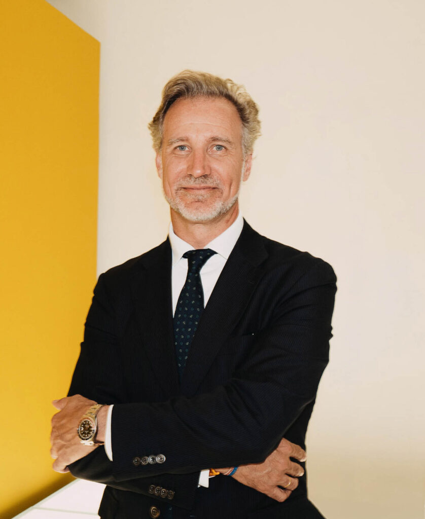 A man in a dark suit and tie stands with arms crossed, wearing a wristwatch, in front of a plain wall with a yellow panel.