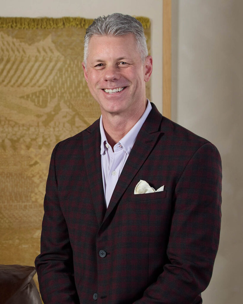 A man with short gray hair, wearing a checked dark blazer, white shirt, and pocket square, stands smiling in front of a patterned wall hanging.