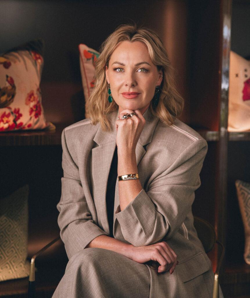 A woman with blonde hair in a beige blazer sits with one hand resting on her chin. Patterned pillows are visible on shelves in the background.