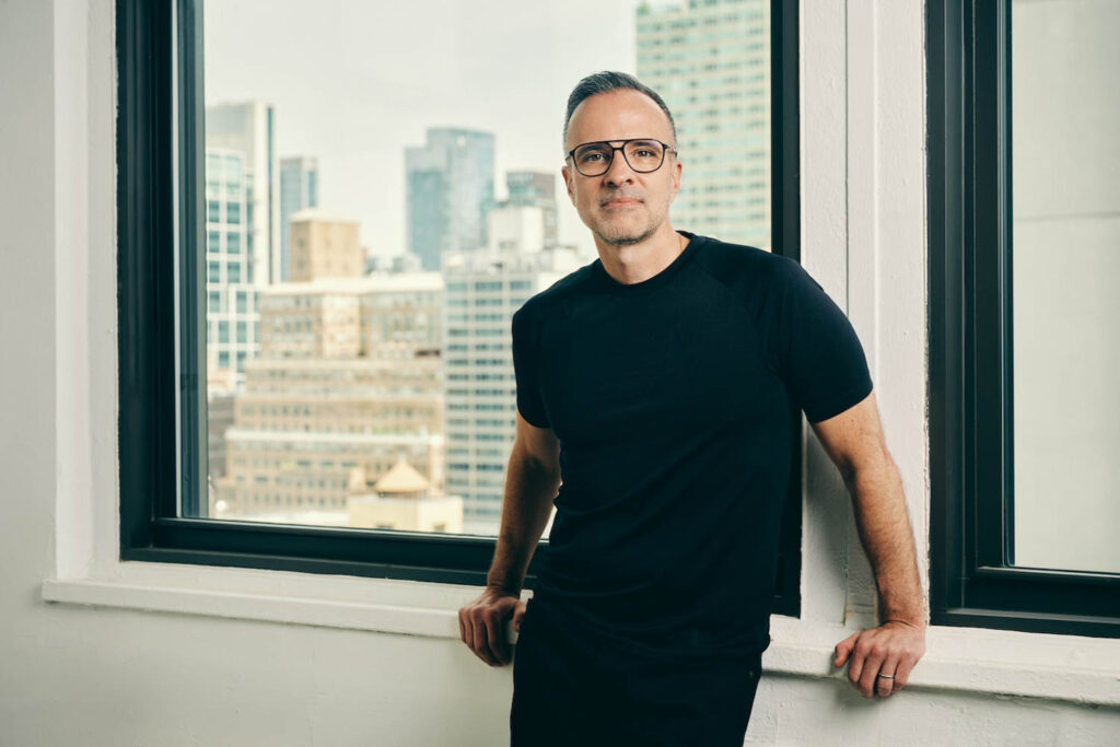 A man wearing glasses and a black t-shirt stands by a windowsill with a cityscape visible through large windows behind him.