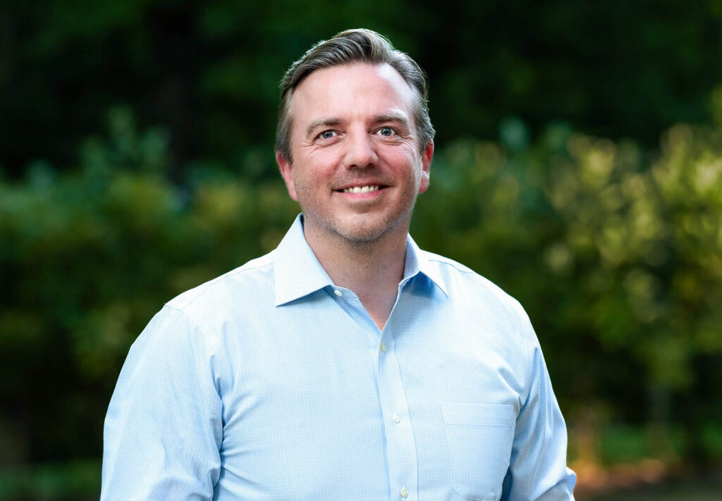 A man in a light blue button-up shirt stands outdoors, smiling, with greenery blurred in the background.
