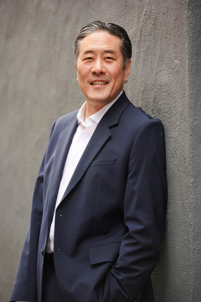 A man in a navy blue suit and white shirt stands against a textured gray wall, smiling at the camera.