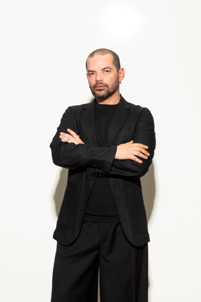 A man with a shaved head and beard stands against a white background, wearing a black jacket, black shirt, and black pants with his arms crossed.
