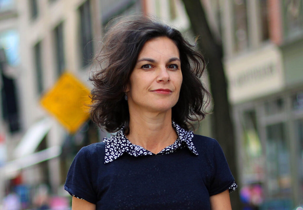 A woman with wavy dark hair stands outdoors in a city, wearing a dark shirt with a patterned collar. Blurred buildings and a yellow street sign are visible in the background.