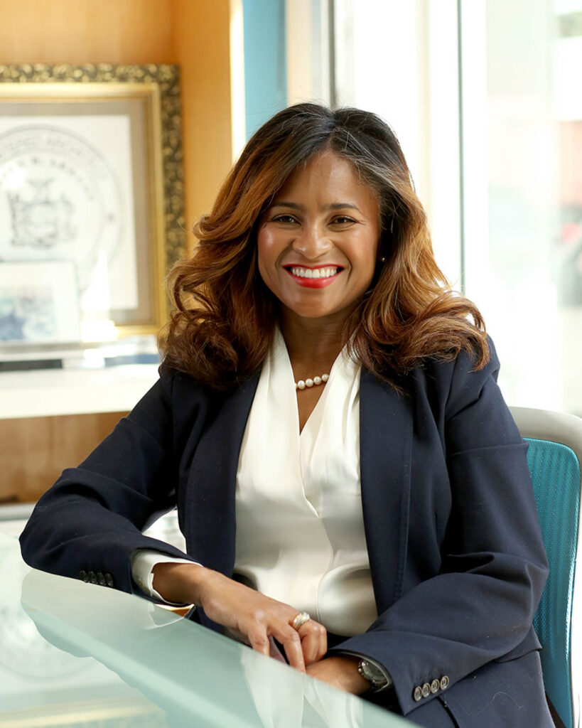 A woman in a navy blazer and white blouse sits at a desk, smiling. Framed documents and photos are visible in the background.