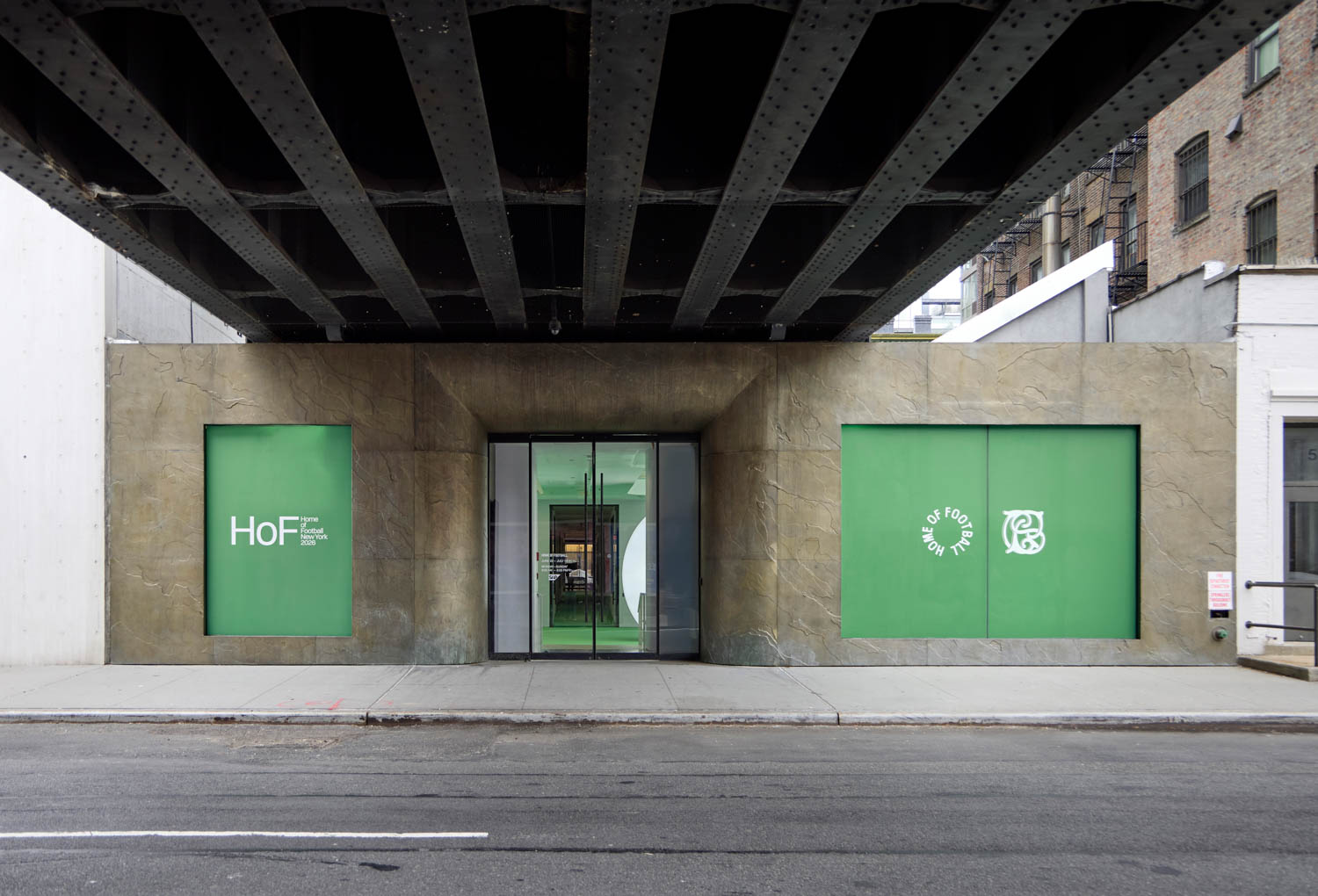 Street view of a modern building entrance with green-framed windows labeled "HoF" and "Home of Food." The structure is beneath a black metal bridge.