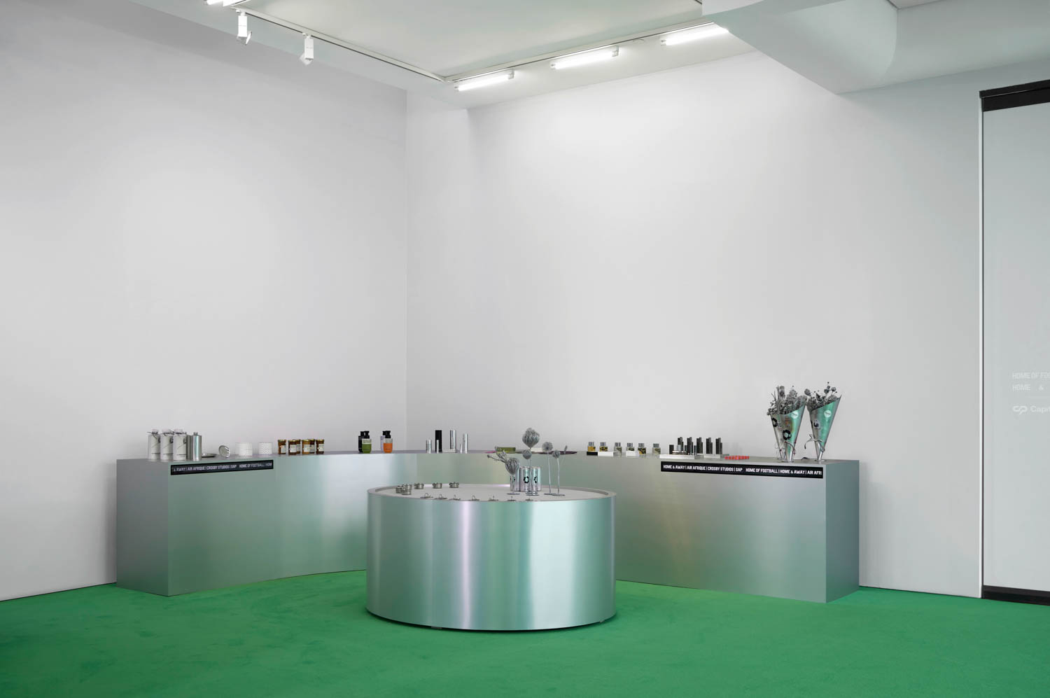 A minimalist retail display features metallic counters with various products arranged on top, set against white walls and a green carpeted floor.