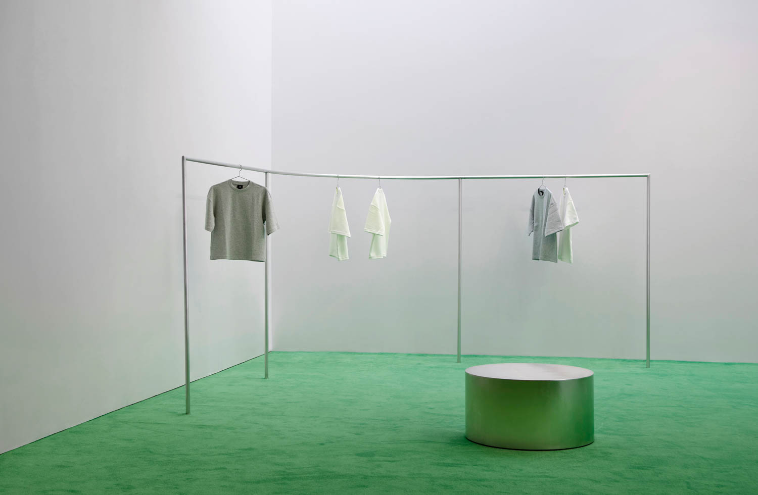 Minimalist room with green carpet, a silver clothes rack displaying neutral-colored garments, and a round metallic stool in the center.
