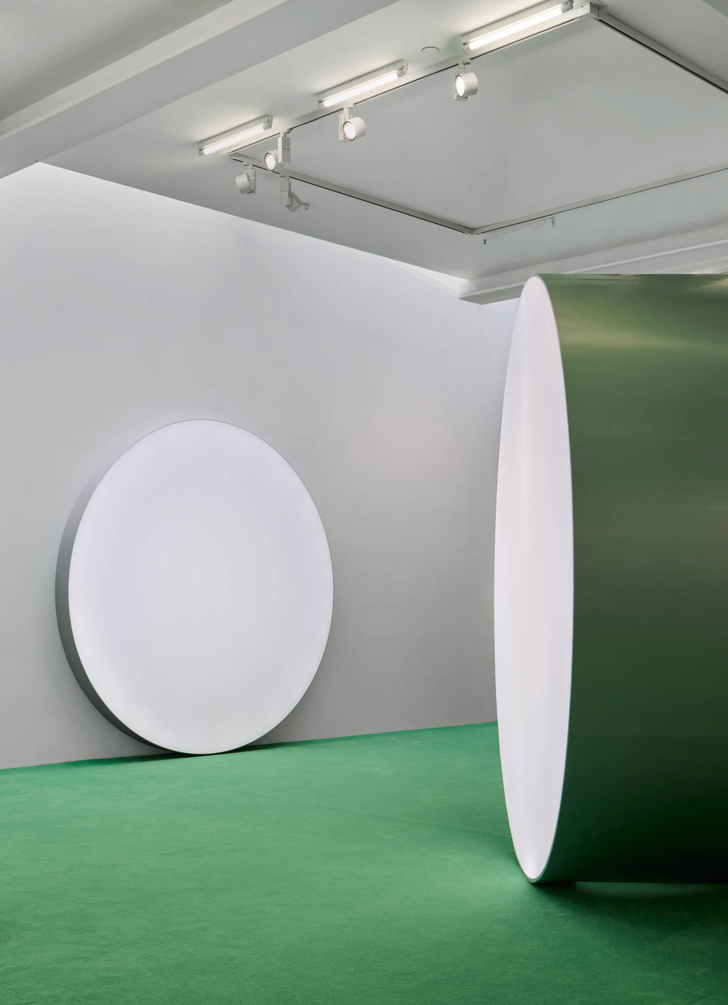 A large white circular object leans against a white wall, while a tall green cylindrical structure stands on green carpet in a minimalist gallery space.