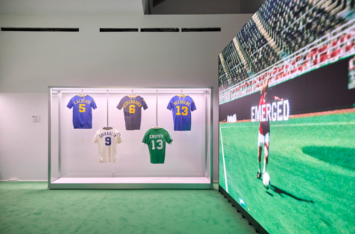 Display case with five vintage soccer jerseys and a large screen showing a soccer match featuring a player running on the field.