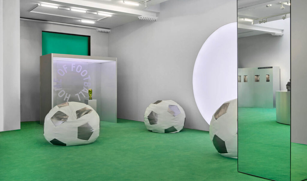 A modern room with green carpet, soccer ball-shaped bean bags, a mirror, a display case with a trophy, and a large circular light on the wall.