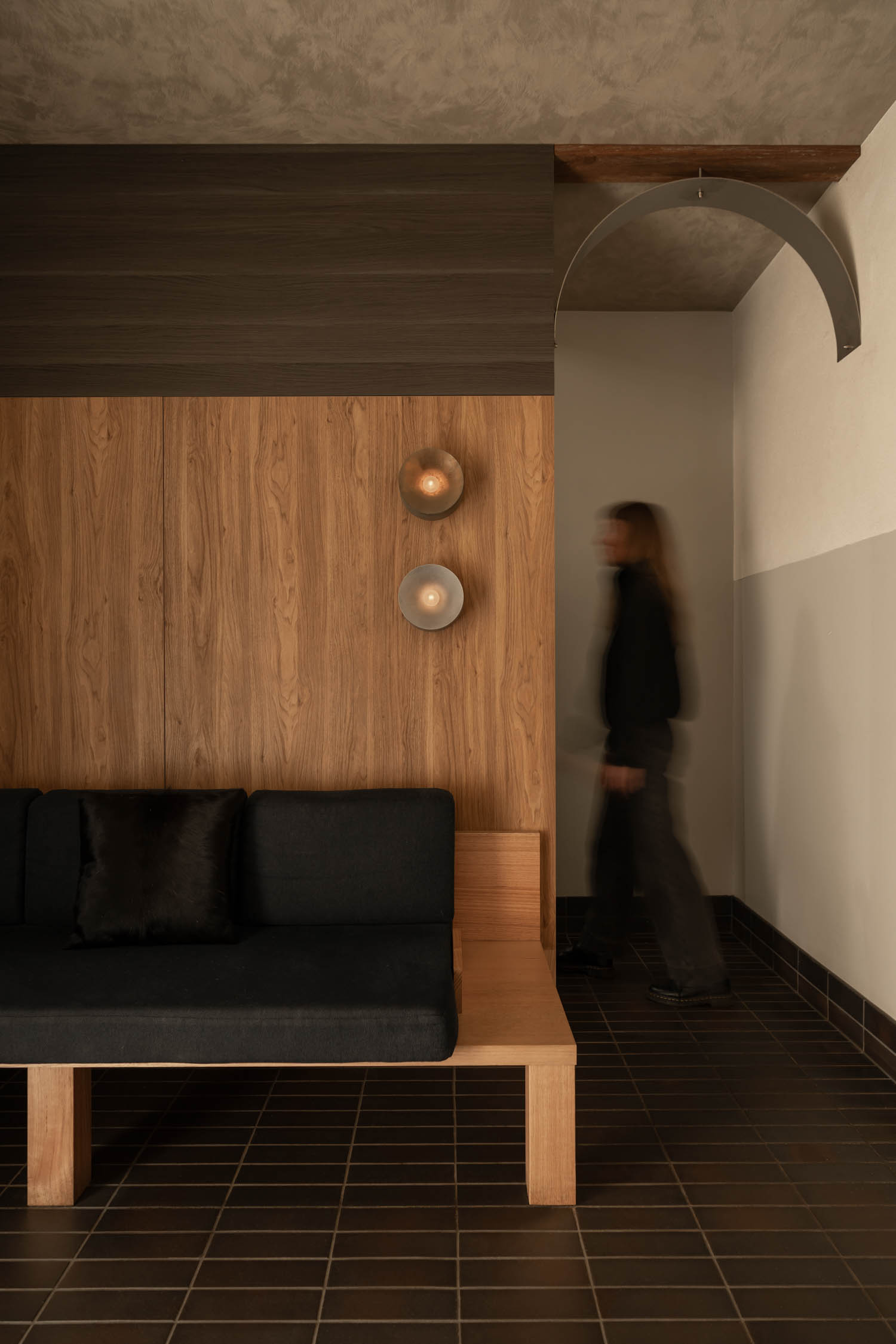 A person walks past a wooden bench with a black cushion in a modern room with wood paneling, wall lights, and tile flooring.