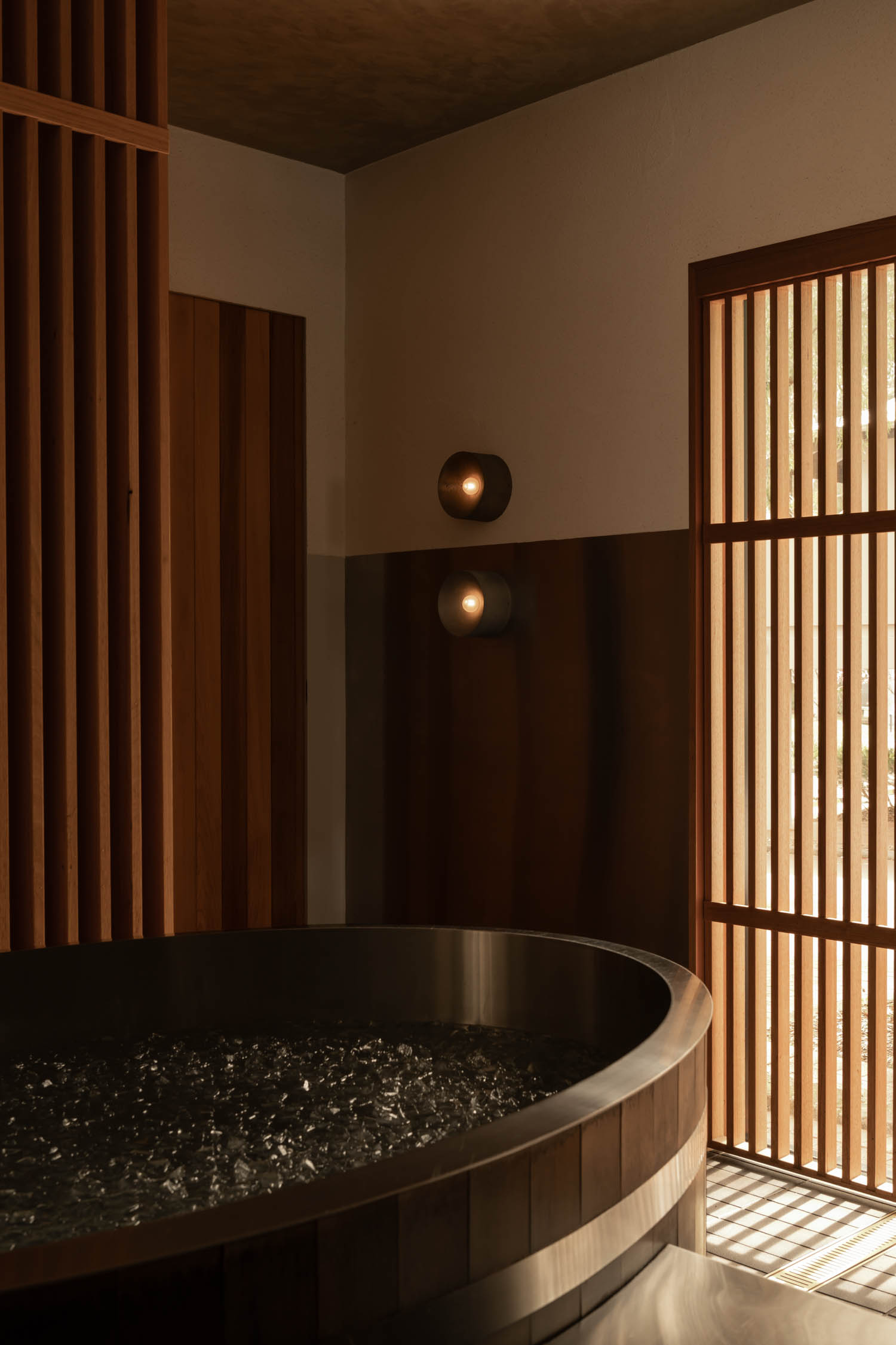 Wood-paneled bathroom with a round soaking tub, wall-mounted lights, and vertical wooden slats allowing natural light into the space.