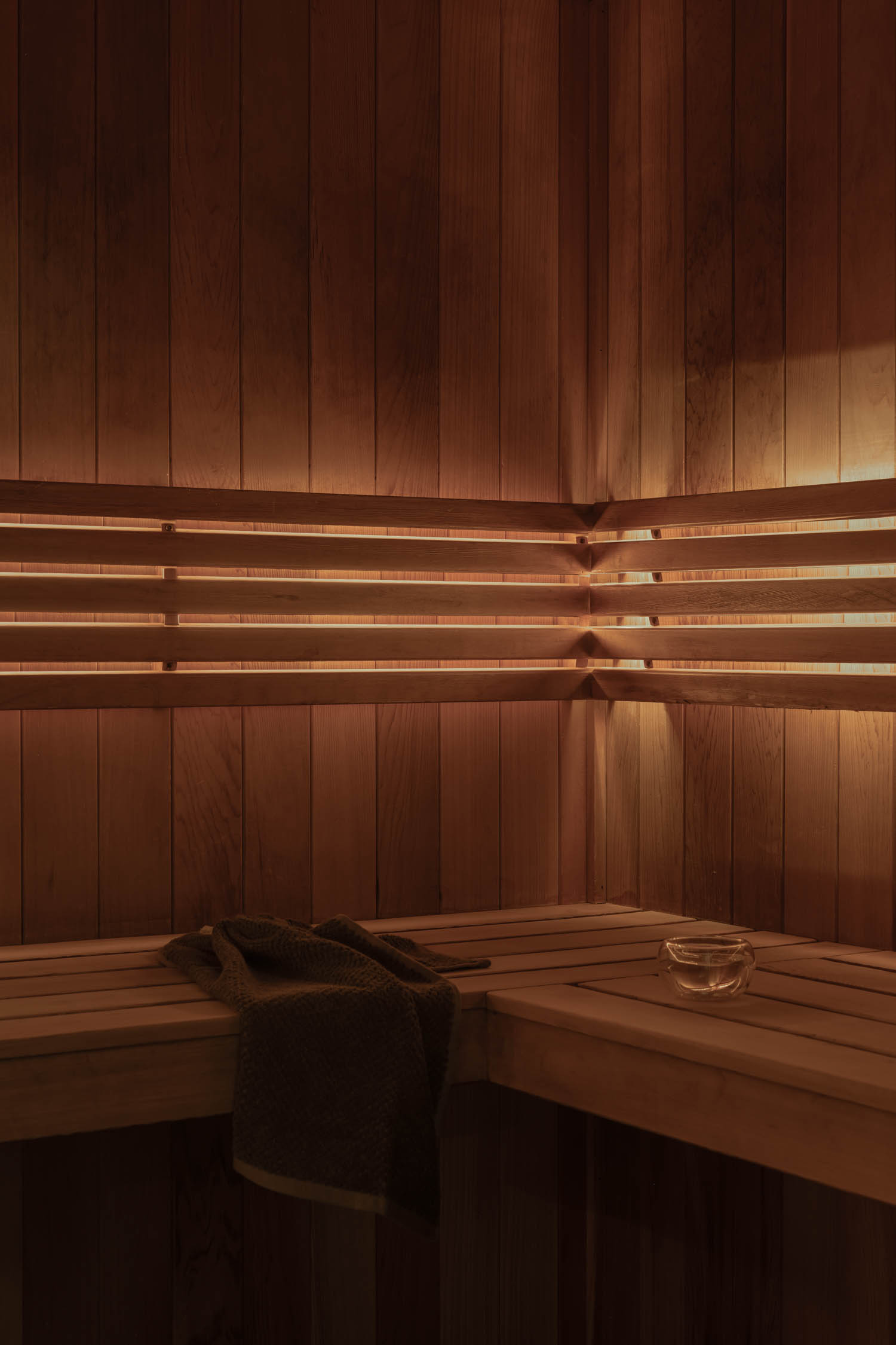 Wood-paneled sauna with built-in bench, illuminated by warm lights along the walls. A towel and a small glass bowl rest on the corner of the bench.