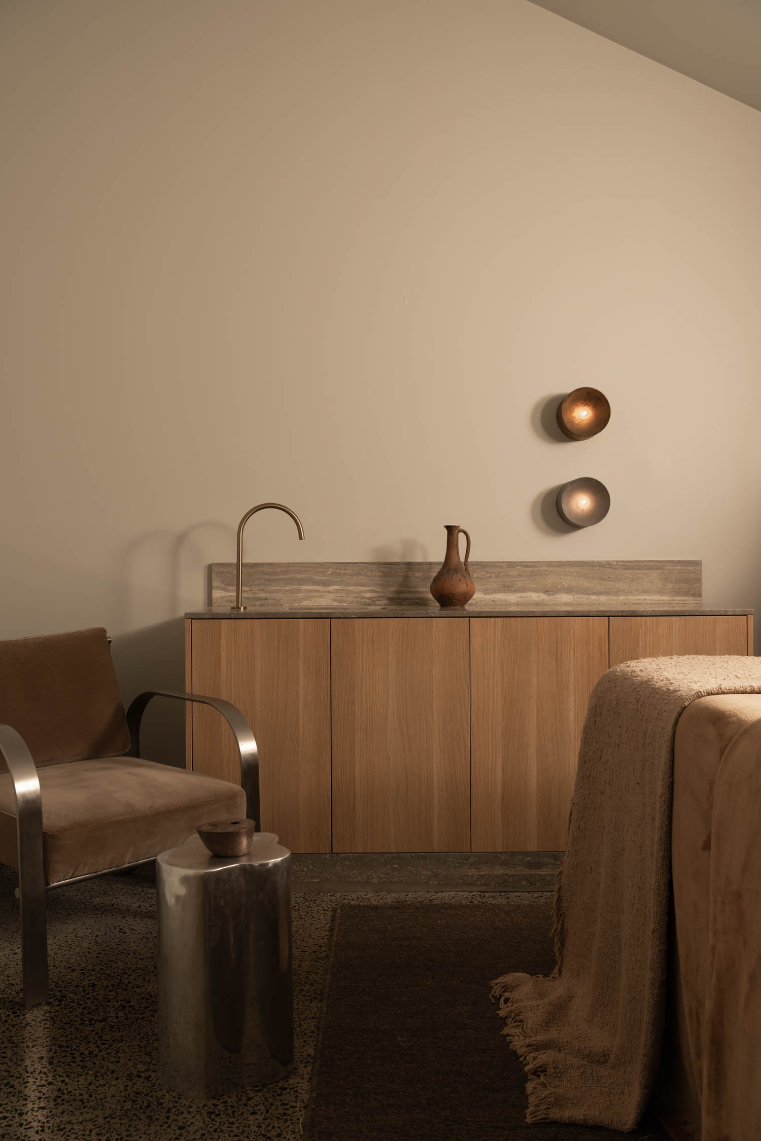 Minimalist room with a tan chair, wood cabinet, metallic sink, ceramic vase, textured blanket, and two round wall lights on a beige wall.