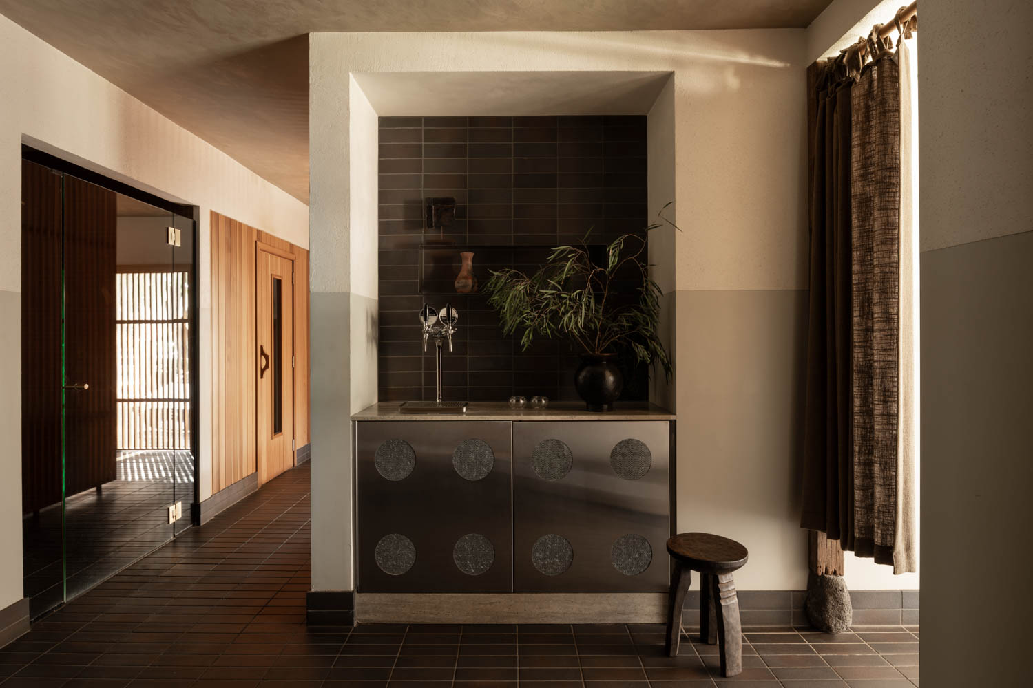 Small kitchenette with dark tiled backsplash, minimalist cabinets, a plant, and decorative objects on the counter; stool and window with brown curtains at the side, hallway visible to the left.