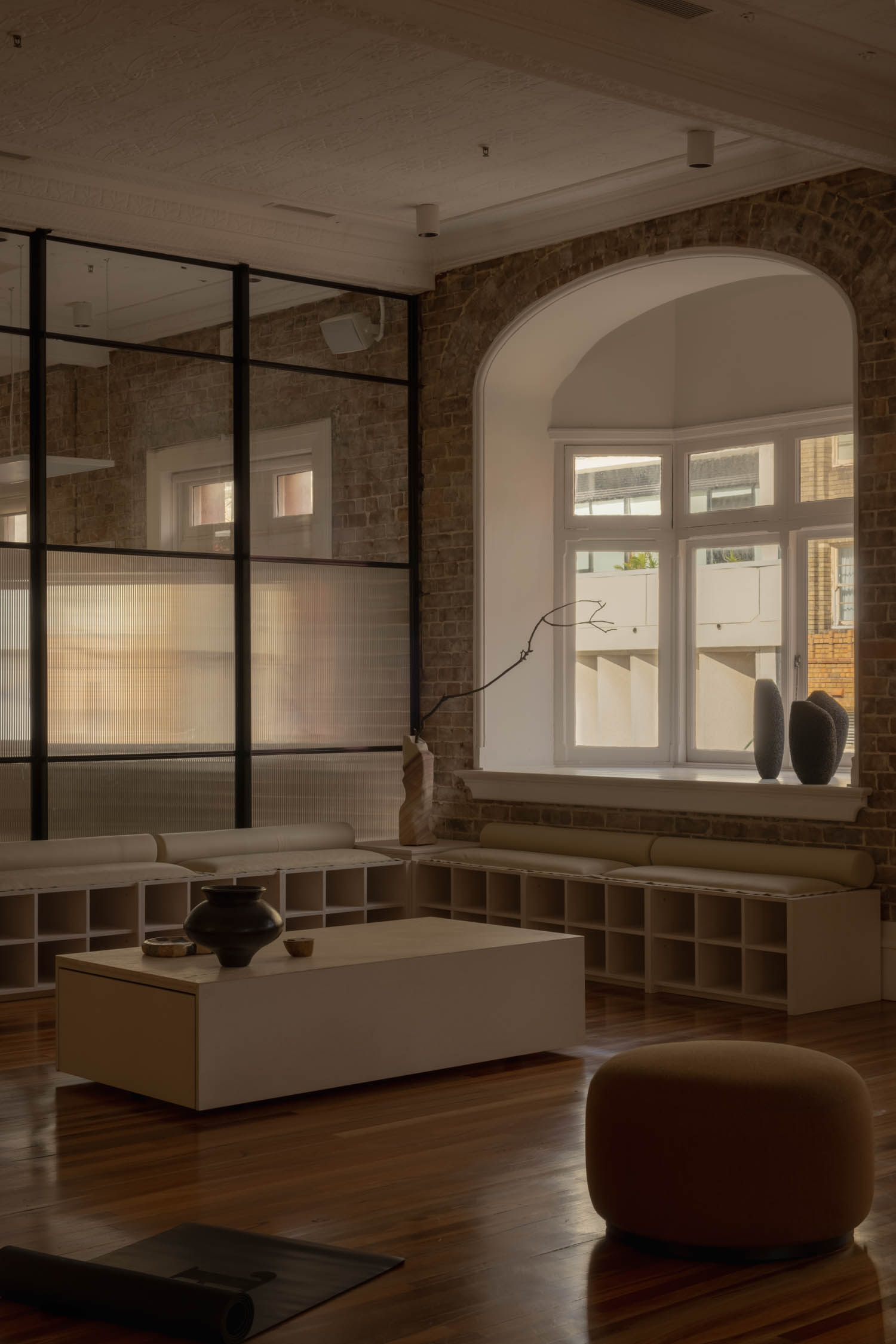 Minimalist living room with brick walls, large arched window, white shelving, neutral-toned furniture, and decorative vases on the windowsill. Natural light fills the space.