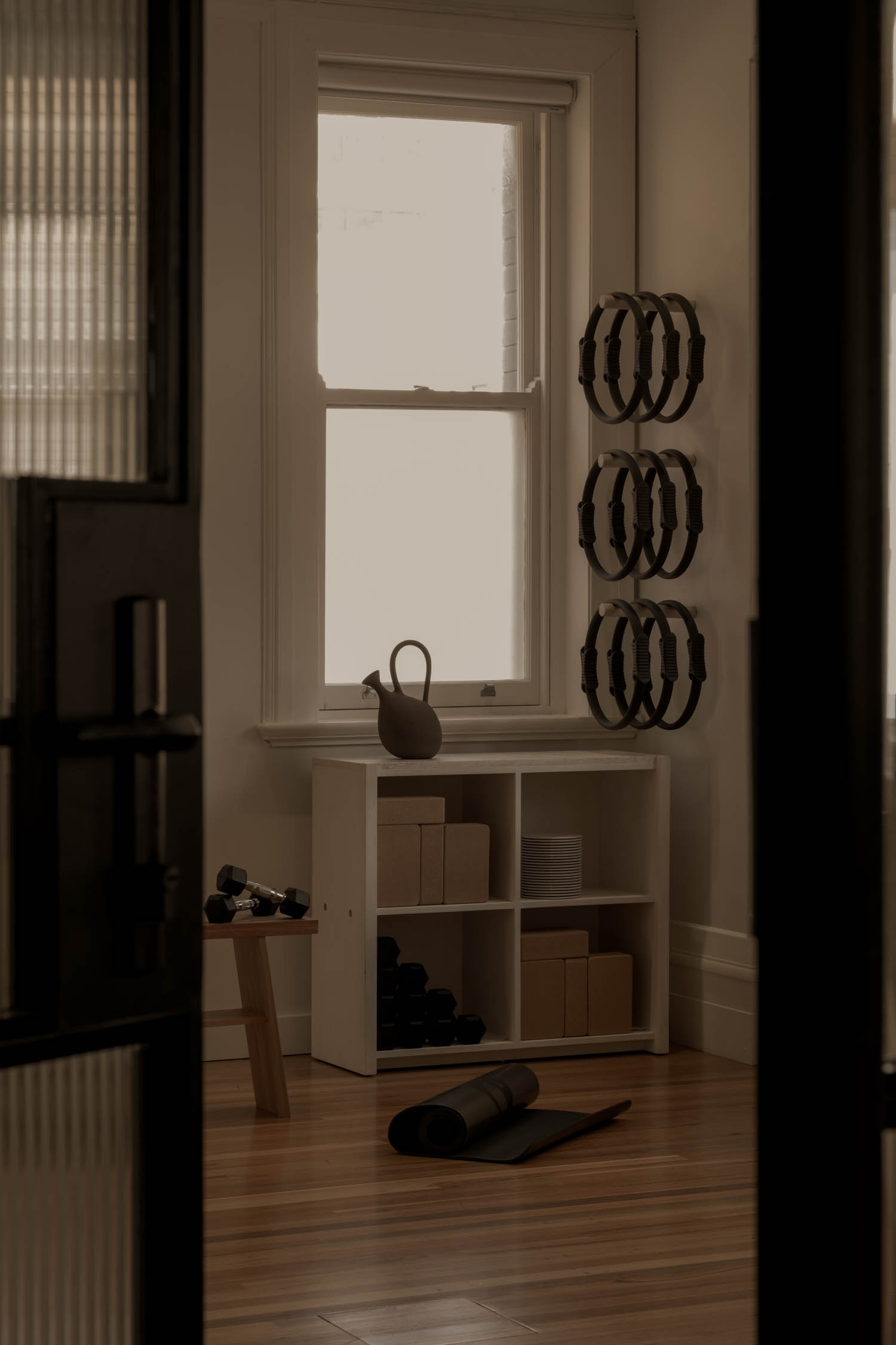 Small home gym with dumbbells on a bench, storage shelves with boxes, a kettlebell, exercise rings on the wall, and a rolled yoga mat on a wood floor near a window.