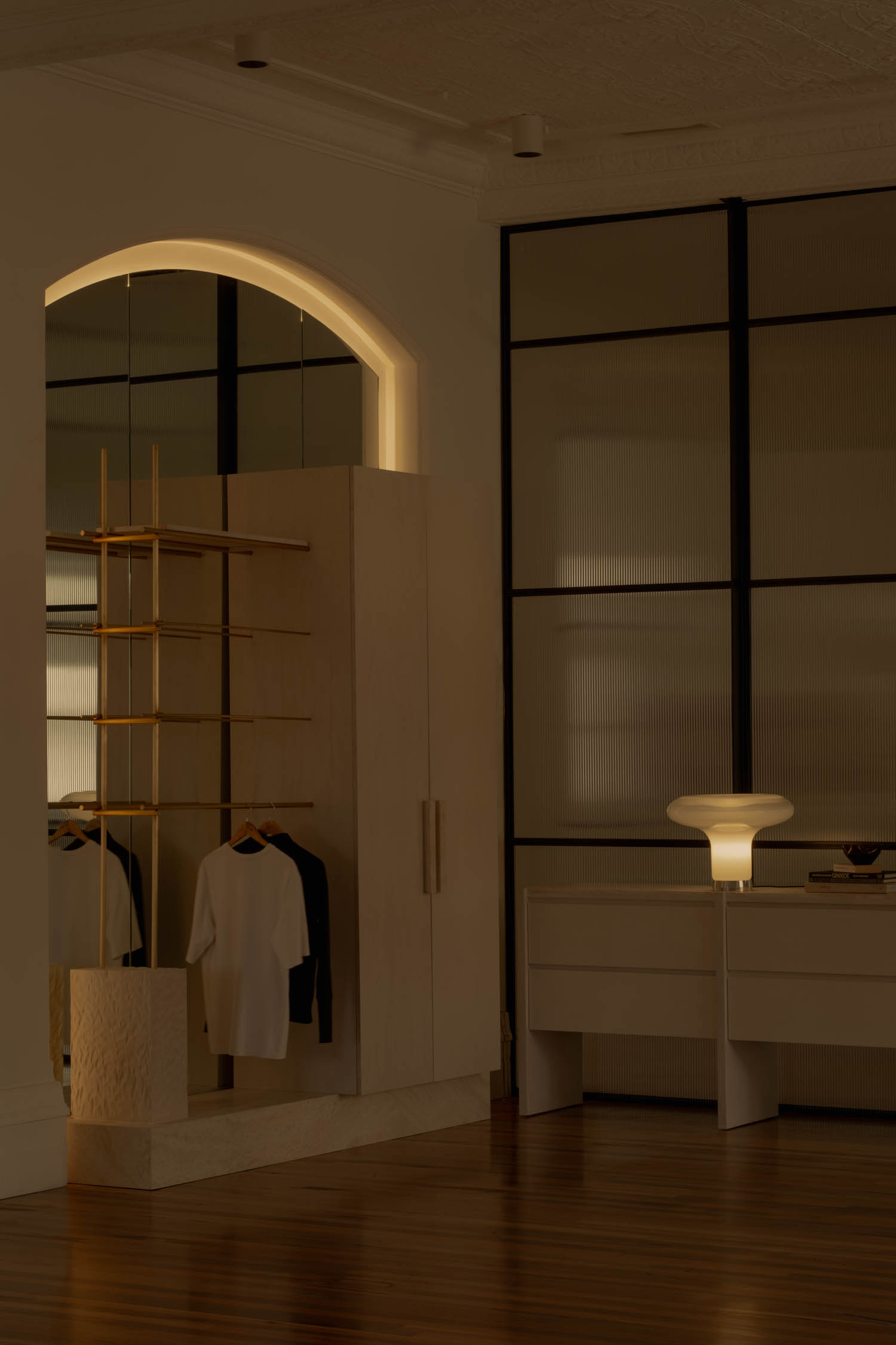 A warmly lit modern closet with hanging clothes, a wooden shelf, and a glowing table lamp on a white dresser against black-framed glass panels.
