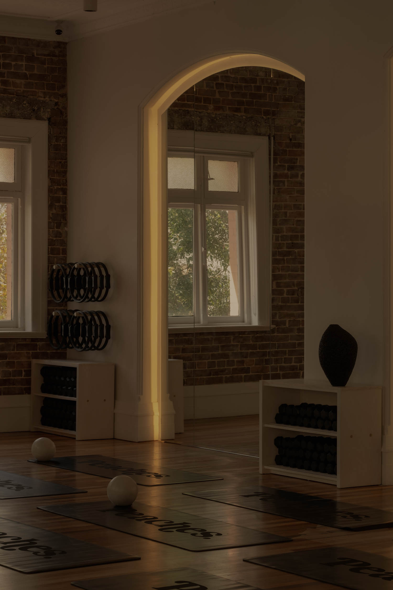 A yoga studio with mats on the wooden floor, shelves holding exercise equipment, and large windows letting in natural light.