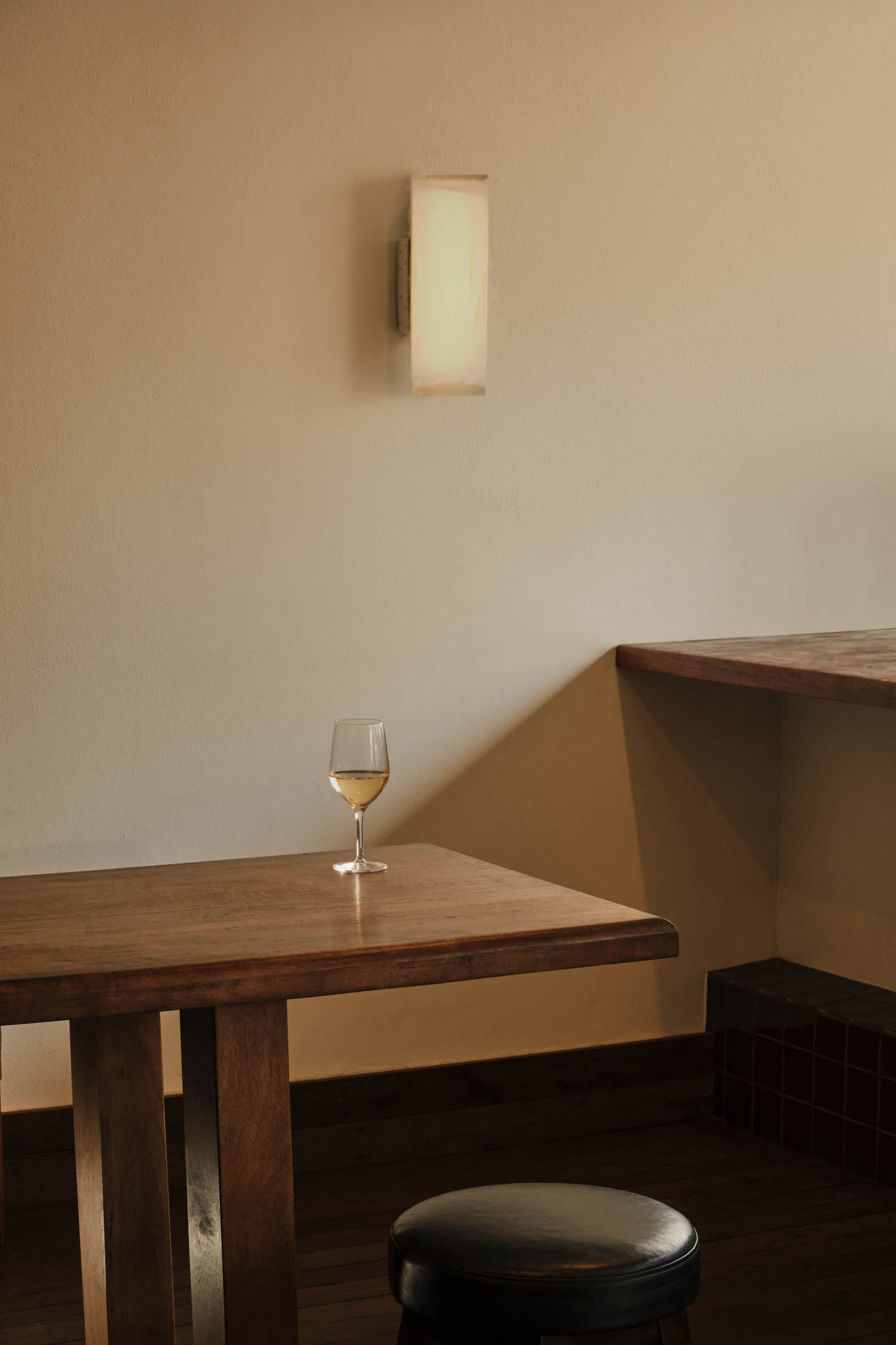 A glass of white wine sits on a wooden table near a wall with a modern sconce; a round black stool is in the foreground.