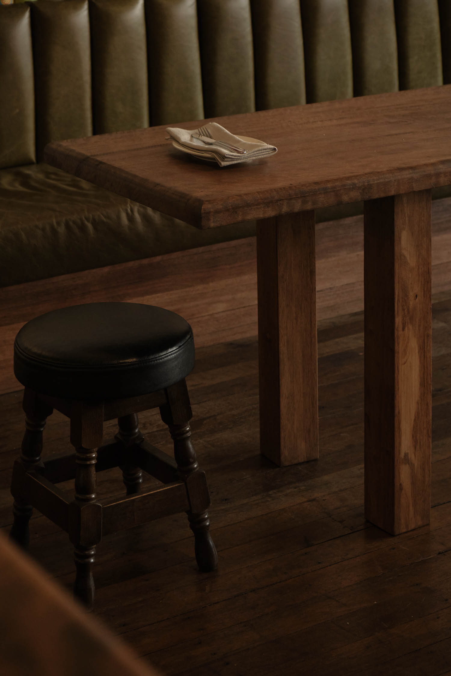 A wooden table with folded napkins and utensils sits next to a round black stool and olive green upholstered bench in a dimly lit room.