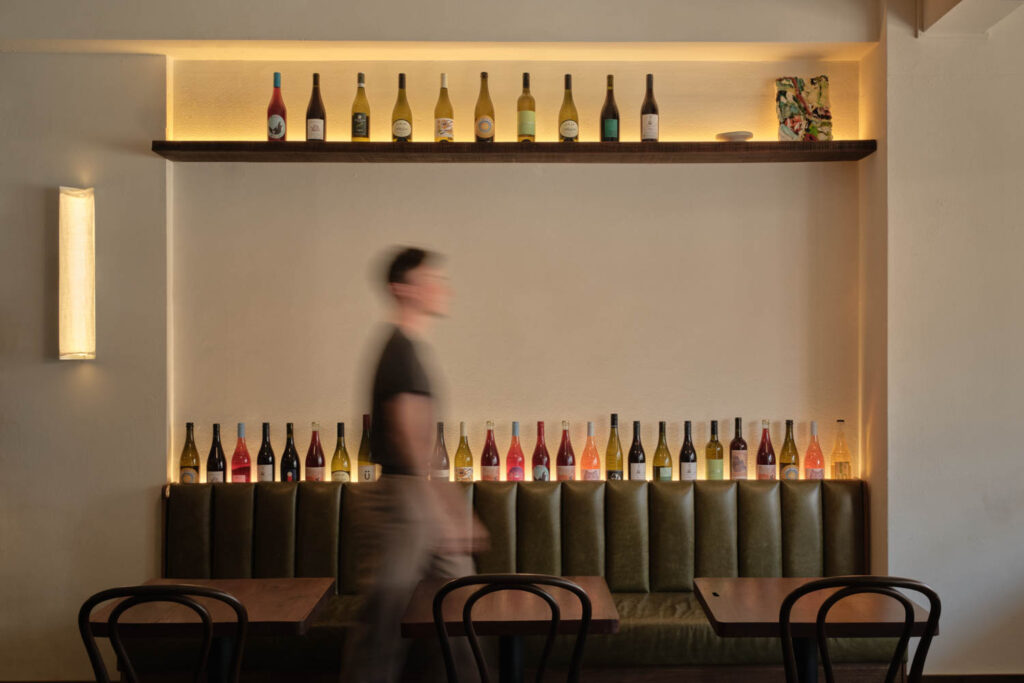 A person walks past a booth with green upholstery, two tables, chairs, and shelves displaying wine bottles on a softly lit wall.