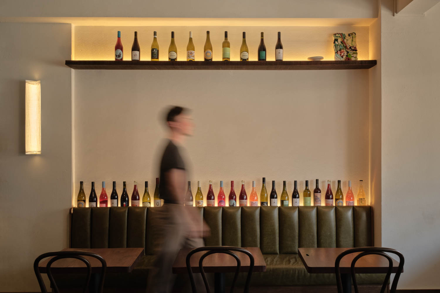 A person walks past a booth with green upholstery, two tables, chairs, and shelves displaying wine bottles on a softly lit wall.