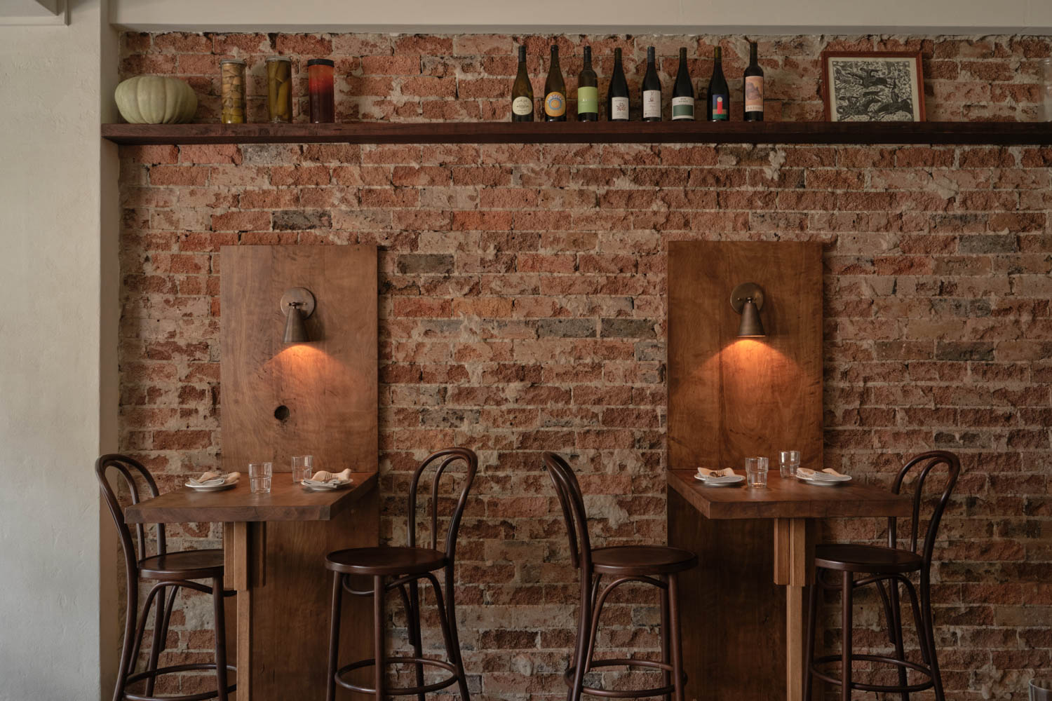 Two wooden tables with chairs sit against a brick wall, each with a wall lamp above. A shelf above displays bottles, jars, and a framed picture.
