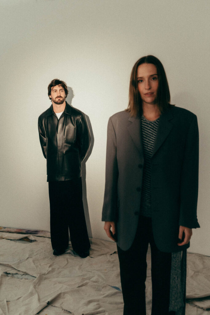 A man in a black leather jacket stands in the background while a woman in an oversized gray blazer stands in the foreground, both indoors on a draped floor.