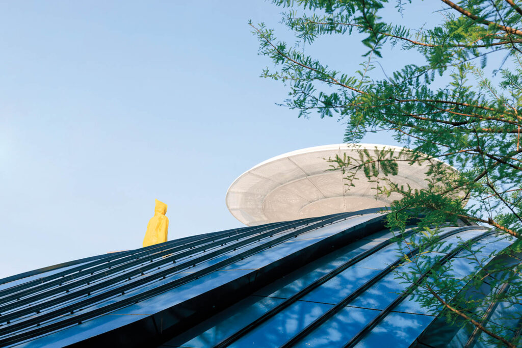 Modern building with a curved glass roof, a white circular structure on top, a yellow statue in the background, and green branches framing the right side against a blue sky.