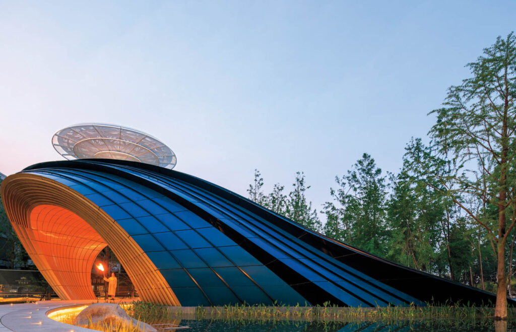 Futuristic building with a curved, blue and black exterior, orange-lit entrance, and circular structure on top, surrounded by trees.