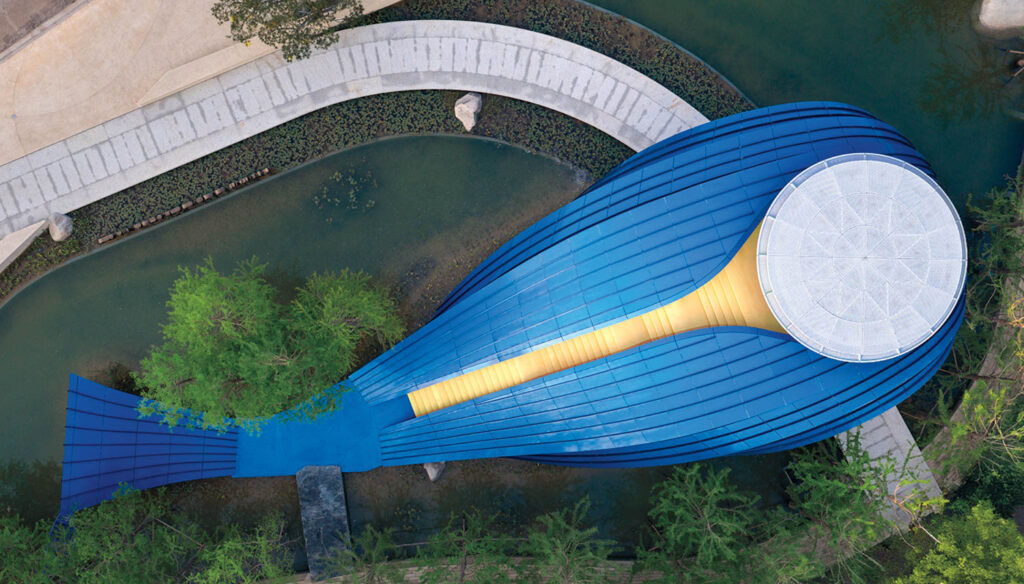 Aerial view of a blue, whale-shaped building with a white circular area at the head, surrounded by water and pathways.