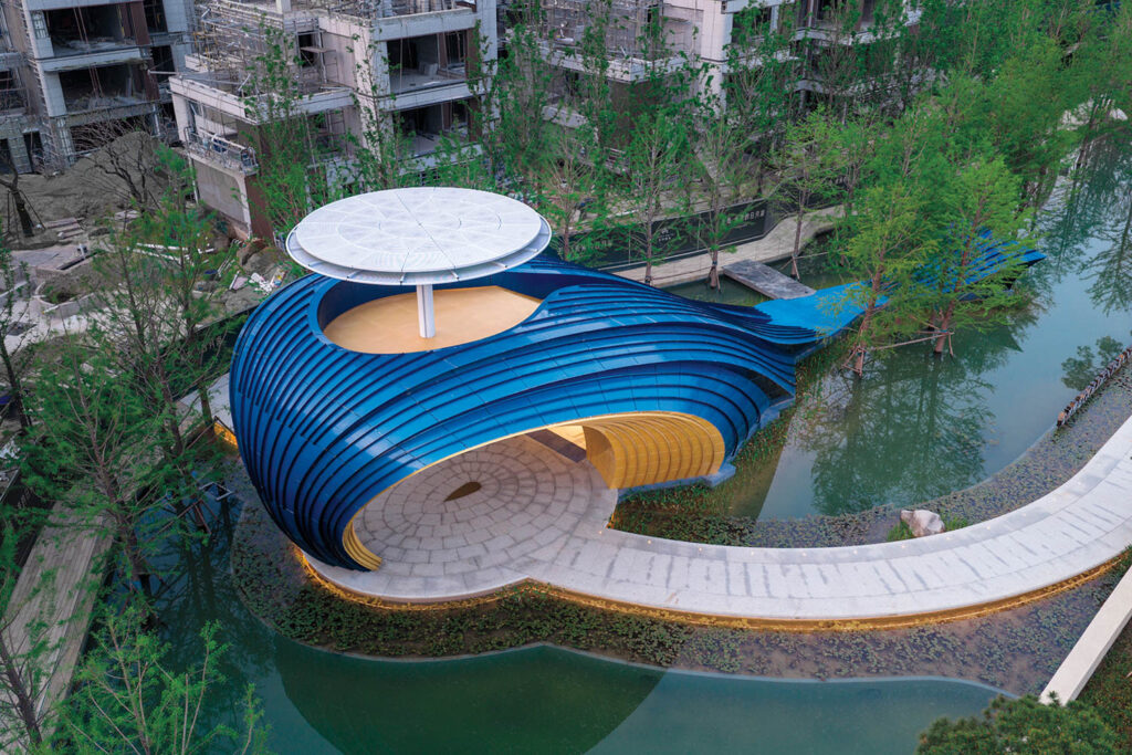 A modern blue and yellow pavilion with curved architecture sits beside a pond, surrounded by trees and unfinished buildings in the background.