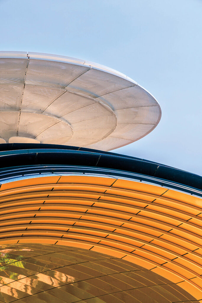 Close-up of two modern architectural structures, one with metallic silver panels above and another with curved golden panels below, set against a clear blue sky.
