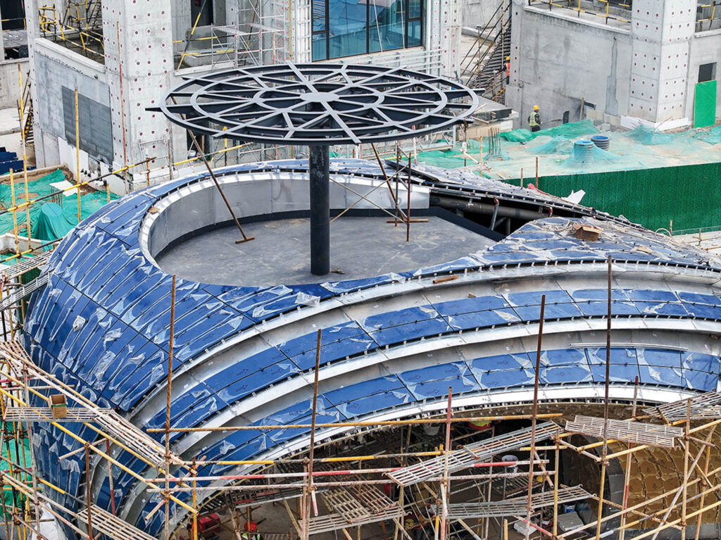 A modern building under construction with scaffolding, blue paneling on a curved roof, and a large circular metal framework on a central pole.