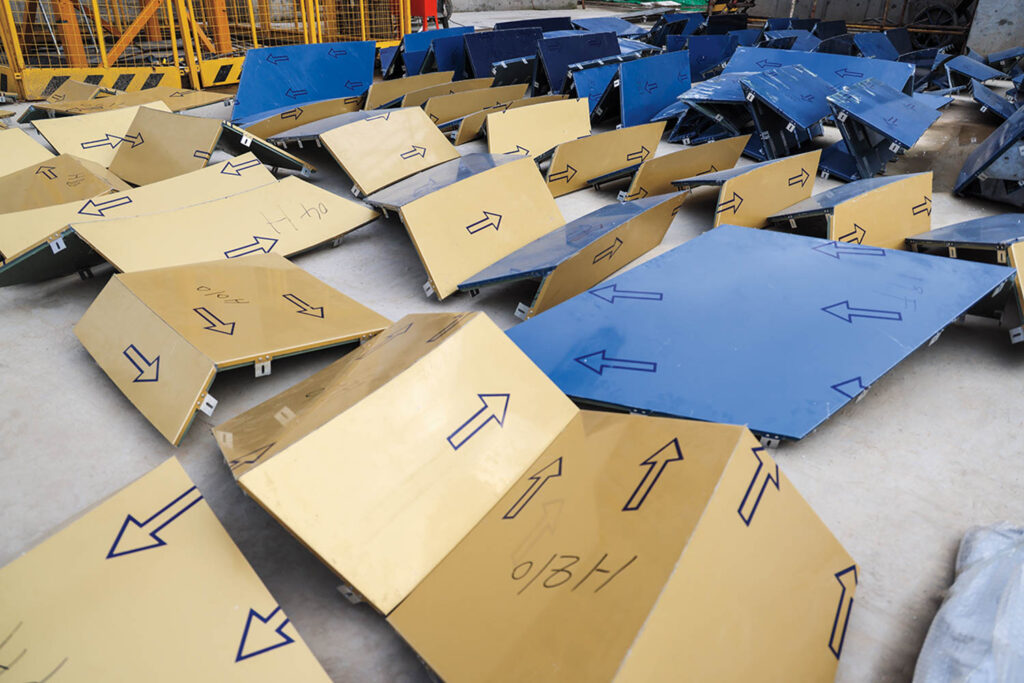 Scattered metal panels with blue and gold surfaces, each marked with arrows and labels, lie on a concrete floor near yellow safety barriers at a construction site.