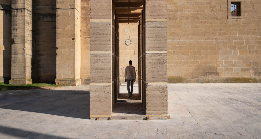 A person stands facing away inside a rectangular stone structure, framed by tall stone walls and an open courtyard under daylight.