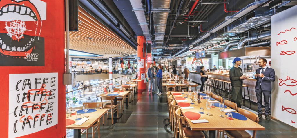Modern restaurant interior with colorful decor, set tables, and two people conversing near the bar; diners and staff are visible in the background under bright lighting.