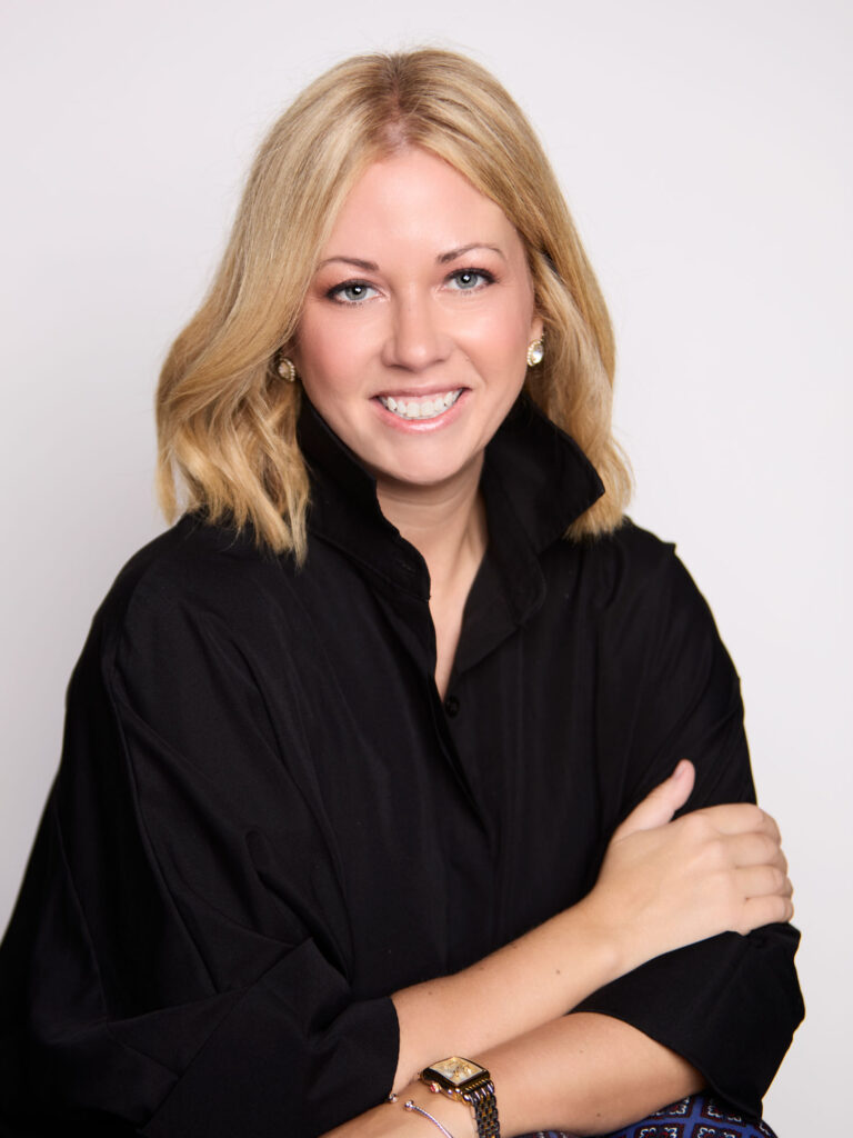 Woman with blonde hair wearing a black shirt, smiling at the camera with her arms folded over each other against a plain background.