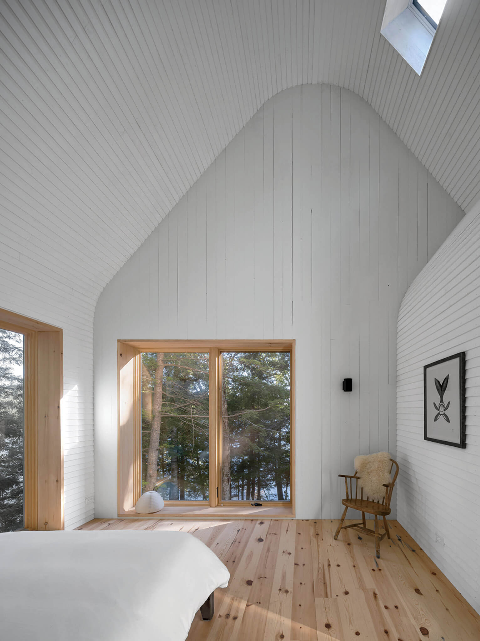 Minimalist bedroom with white walls and wooden floor, large window overlooking trees, skylight above, wooden chair with fur throw, and framed art on the wall.