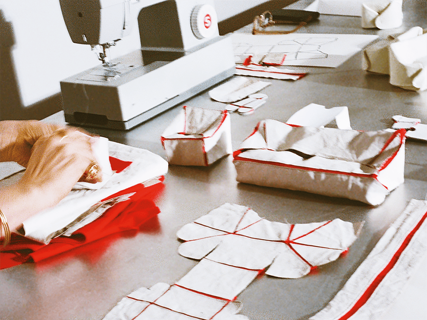 A person sews fabric pieces with red edges at a table, surrounded by cut fabric shapes and a sewing machine.