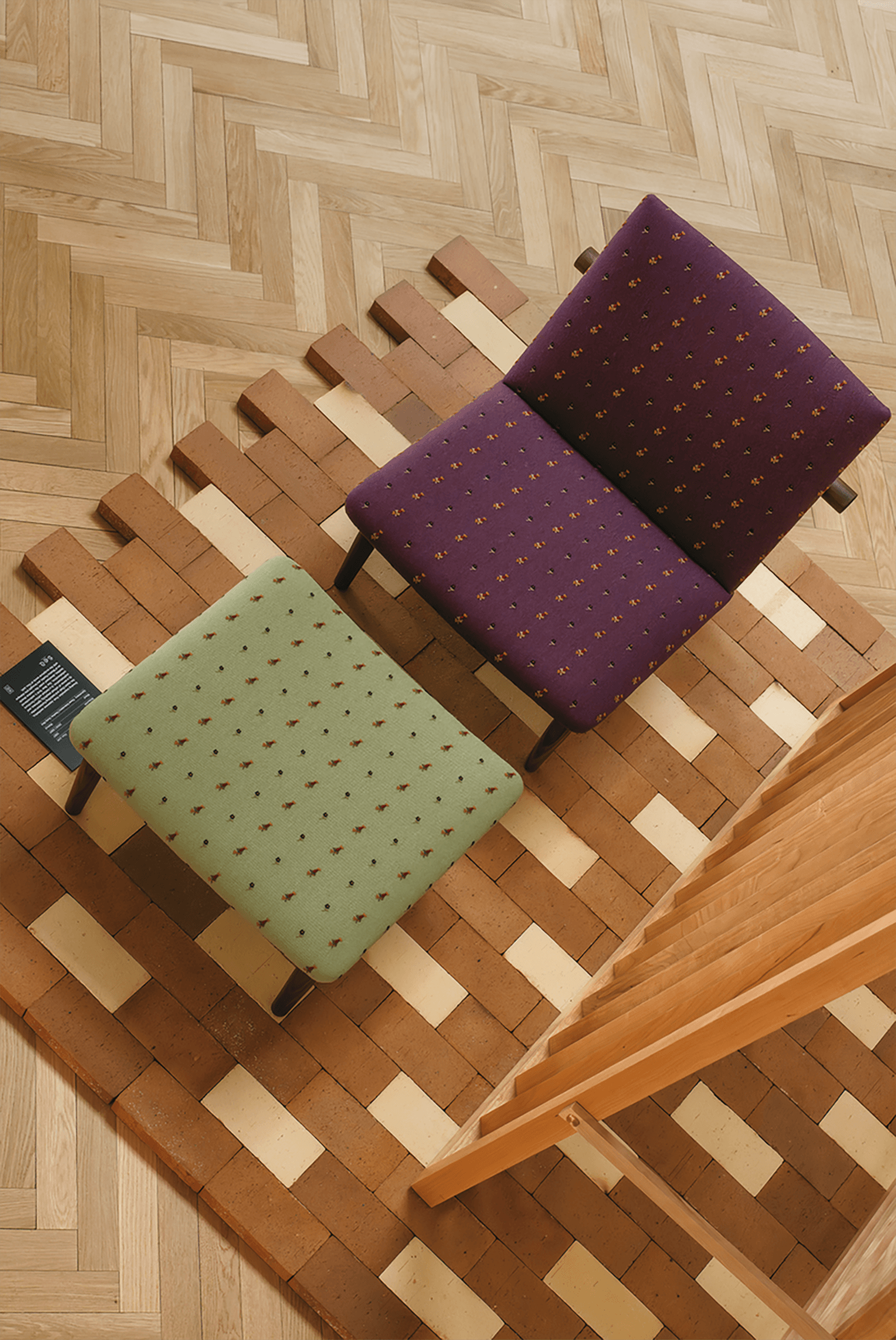 Two chairs, one purple and one green, with dotted upholstery on a woven brick and tile patterned rug, are arranged next to a wooden structure on a herringbone wood floor.